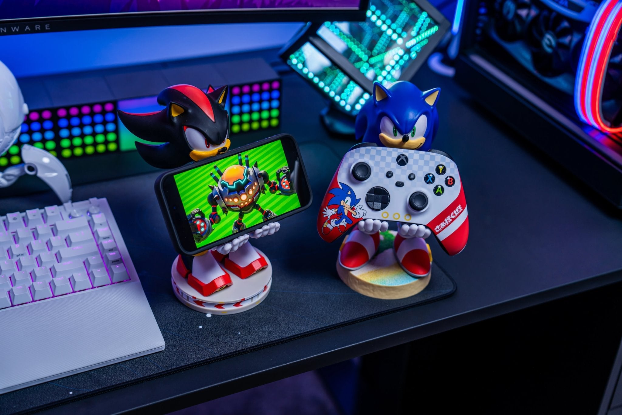 Shadow the Hedgehog and Sonic the Hedgehog Cable Guys displayed on a gaming desk, holding a smartphone and an Xbox controller